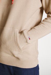 Sudadera con capucha beige con bolsillo frontal, puños y dobladillo acanalados. Incluye cordones rojos y un pequeño logo bordado en la muñeca.