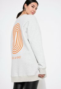 Felpa grigia con vestibilità oversized, caratterizzata da un audace design a cerchi concentrici arancioni e dalla scritta "COOL & KIND" sul retro. Materiale morbido.