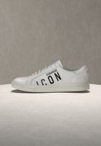 Baskets basses blanches avec texte noir "DSQUARED2 ICON" sur le côté et lacets blancs, placées sur une surface beige neutre et un fond.
