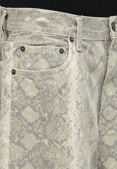 Κοντινό πλάνο στα μπεζ και γκρι τζιν με σχέδιο φίδιού, που δείχνει την μπροστινή τσέπη, τις θηλιές της ζώνης, τα καρφιά και τη λεπτομέρεια του κουμπιού.