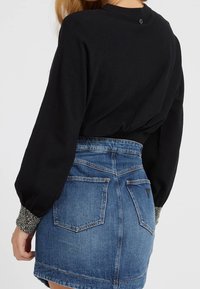 Zwarte trui met volumineuze mouwen, geribbelde manchetten met zilveren versieringen, gecombineerd met een blauwe denim rok met achterzakken.