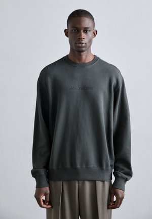 Dark green oversized sweatshirt in cotton blend, crew neck, ribbed cuffs, featuring embossed logo detail on the front, worn over beige trousers.