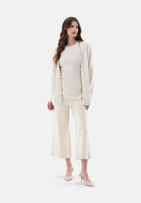 Cremfarbener Cardigan mit großen Knöpfen über einem figurbetonten Top, kombiniert mit weiten Hosen in passendem Farbton und spitzen Pumps. Glatte Textur.