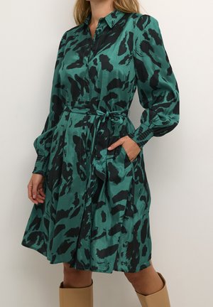 Femme portant une robe à manches longues imprimé animalier vert et noir avec une ceinture à la taille et des poignets froncés, associée à des bottes hautes couleur camel.