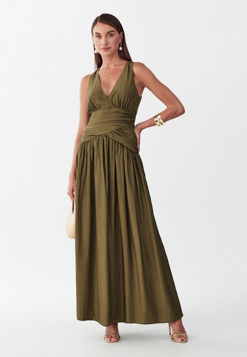 Vrouw draagt een mouwloze olijfgroene maxi-jurk met V-hals, houdt een beige gevlochten tas vast, gouden armband, oorbellen en beige sandalen met hoge hakken.