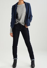 Blazer azul marino sobre un suéter gris, jeans oscuros con los puños doblados, botines negros. Tejido suave con un ajuste casual y sin patrones visibles.