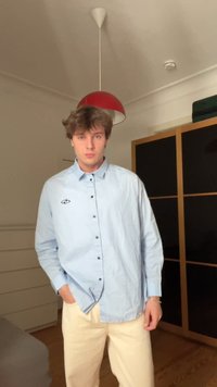 Világoskék gombos ing gallérral, fekete gombokkal, bal mellkason logóval, krémszínű nadrággal viselve, beltérben, fa padlón.