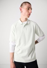 Lichtgroen polo-shirt met een kraag, korte mouwen en subtiele branding. Gedragen over een wit langemouwen shirt.