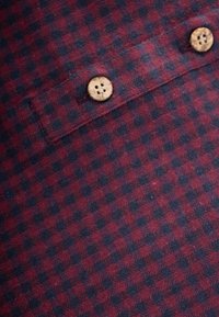 Tessuto a quadri in un profondo rosso e blu navy, con una trama testurizzata, con due bottoni in legno visibili su una patta della tasca.