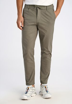 Pantalones cónicos color caqui con cintura ajustable, dos bolsillos laterales y una textura sutil. Combinado con zapatillas blancas con acentos de color.