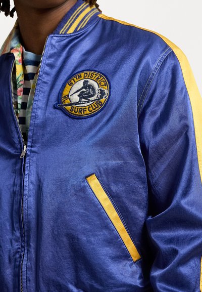 Blaues Satin-Bomberjackett mit gelben Streifen und einem Surfclub-Patch. Verfügt über einen Frontreißverschluss und seitliche Taschen. Glatte Textur.