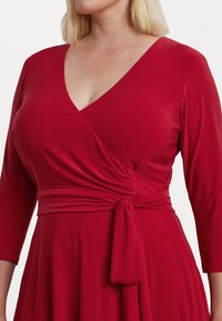 Vestido rojo ajustado con un escote en V profundo, mangas tres cuartos y detalle de cintura atada. Fabricado con una tela suave y ligera.