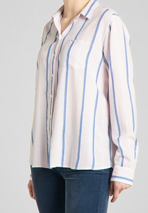 Femme portant une chemise boutonnée à manches longues avec des rayures verticales bleues et blanches, associée à un pantalon bleu foncé.