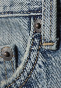Light blue denim fabric featuring contrasting blue stitching, two copper buttons, and a frayed edge near the pocket area.