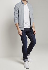 Sudadera gris con cremallera y cuello alto sobre una camiseta blanca, combinada con jeans azul oscuro y zapatillas blancas, con los puños doblados.