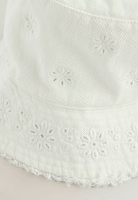 Hvid bøttehat lavet af tekstureret stof, med broderede blomstermønstre langs kanten og en frynset kant.