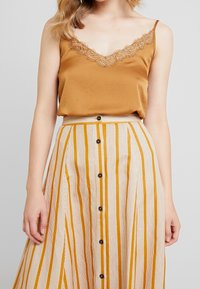 Top de tirantes en mostaza con detalles de encaje, combinado con una falda en línea A a rayas en crema y ámbar, que cuenta con botones en el frente y una cinturilla.