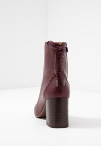 Botín de cuero burdeos hasta el tobillo con un alto tacón cuadrado, que cuenta con un detalle de cordones en la parte posterior y acentos metálicos. Textura suave con un diseño elegante.