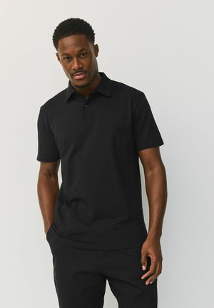 Mees mustas lühikese varrukaga polo-särgis ja mustades pükstes seisab ühe käega taskus tavapärasel heledal taustal.