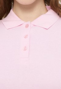 Φωτεινό ροζ πόλο μπλουζάκι από ριμπ ύφανση, με κλασικό γιακά και τέσσερα κουμπιά στο ίδιο χρώμα κατά μήκος της λαιμόκοψης για κλείσιμο.