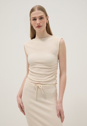Vrouw met stijl blond haar die gouden creolen draagt en een gestructureerde crèmekleurige mouwloze top en bijpassende rok met trekkoord in de taille.