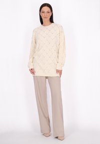 Cremfarbener gestrickter Pullover mit Rautenmuster, kombiniert mit hellbeigen, weitem Hosen und neutralen Absätzen. Glattes, strukturiertes Material.