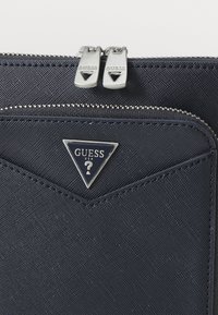 Mørkeblå teksturert syntetisk materiale lommebok med to sølvglidelåser og en trekantet logo plate med "GUESS USA" i hvitt.