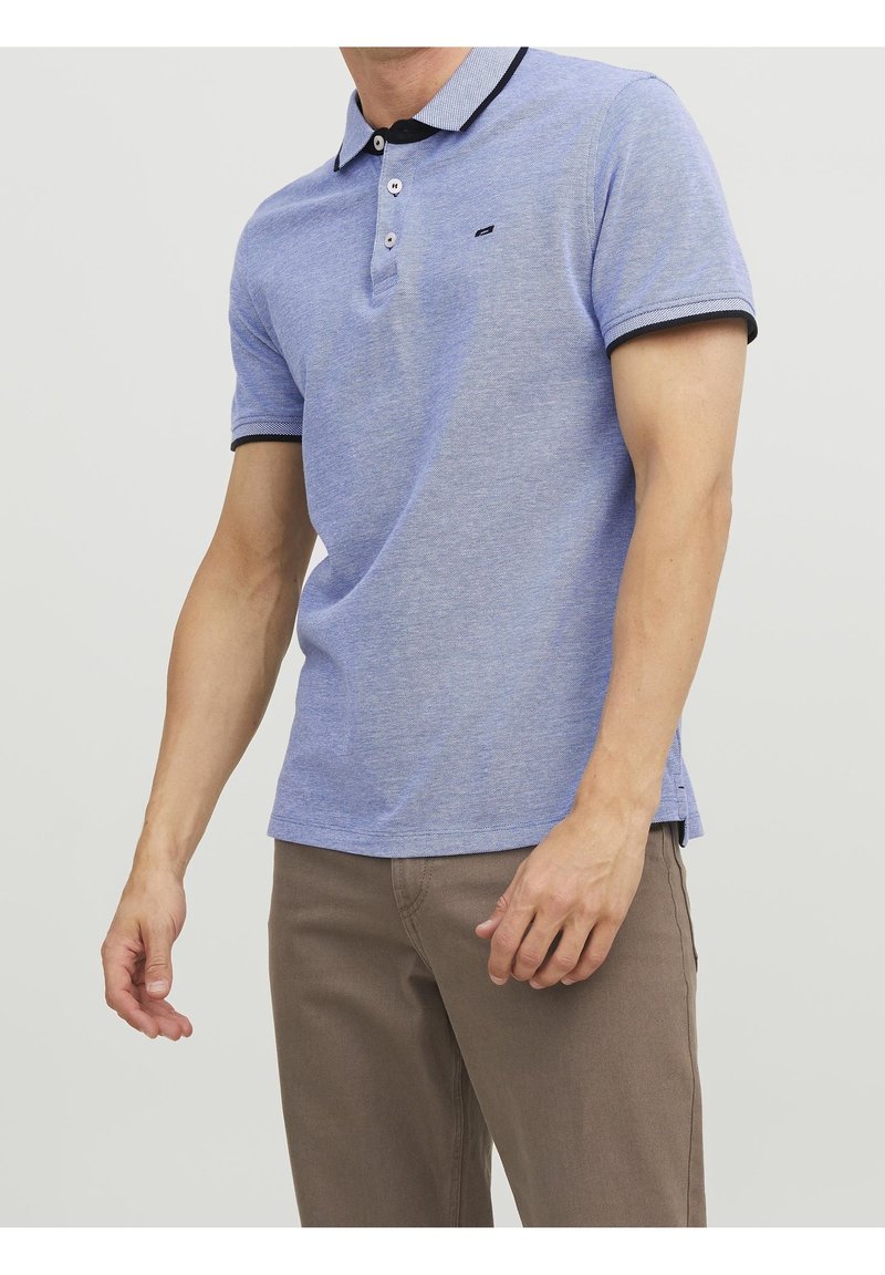 Homme portant un polo à manches courtes bleu clair avec bordure noire et un pantalon beige, debout devant un fond blanc uni.