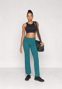 Μαύρο crop top, πετρόλ παντελόνι με φαρδιά πόδια και μαύρα παπούτσια. Το μοντέλο κρατάει μια μαύρη τσάντα duffel. Λείο υλικό, εφαρμοστή σχεδίαση, χωρίς ορατά σχέδια.