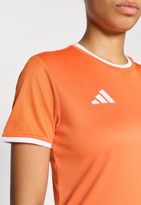 Πορτοκαλί αθλητική φανέλα με κοντά μανίκια, λευκή λεπτομέρεια στο γιακά και λογότυπο με τρεις λευκές ρίγες στην αριστερή πλευρά. Λείο, αεριζόμενο ύφασμα.