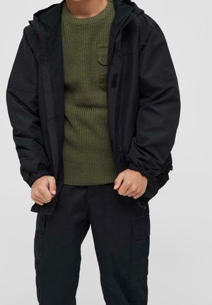 Person iført en sort hættetrøje over en olivengrøn striktrøje med frontlomme, kombineret med sorte cargobukser, foran en grå baggrund.