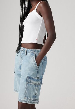 Kvinde med lange fletninger iført en hvid ærmeløs crop top og lyseblå denim cargo shorts med lommer, hånd i lommen, på grå baggrund.