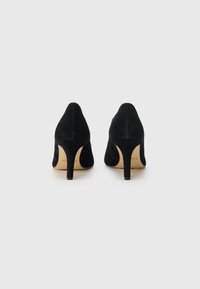 Mustat mokka-korkokengät, joissa on kaareva siluetti, teräväkärkinen muoto ja kapea stiletto-korko. Sisäpohjassa on beige väri logotiedoilla.