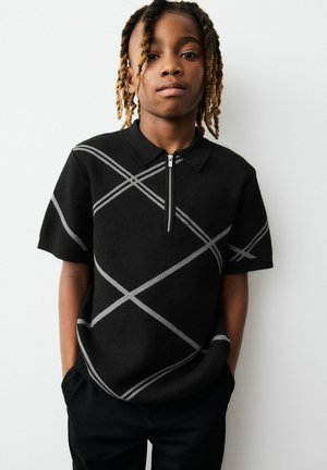 Joven con cabello trenzado que lleva una camiseta polo negra de manga corta con rayas diagonales grises y cremallera frontal, manos en los bolsillos.