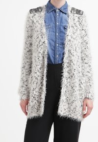 Vit fluffig cardigan med svarta pricker, öppen frontdesign och metalliska axelaccenter, tillsammans med en denimskjorta under.