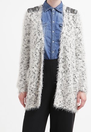 Cardigan bianco soffice con macchie nere, design aperto fronte, e dettagli metallici sulle spalle, abbinato a una camicia di denim sotto.