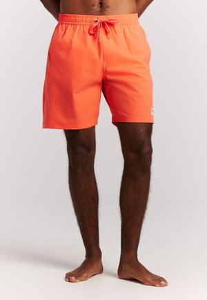 Man die feloranje zwembroek draagt met elastische tailleband en trekkoord, blootsvoets staand tegen een witte achtergrond.