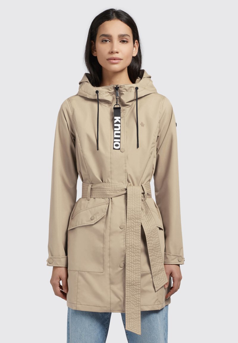 Beige rain jacket with a hood, elastic drawstrings, belted waist, and multiple front pockets. Features a zipper and contrasting label.