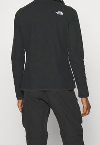 Svart fleecejacka med långa ärmar, figurnära design och en liten vit logotyp på övre ryggen. Slät textur, minimalistisk stil.