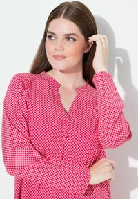 Czerwona bluzka w wzory z lekkiego materiału, z dekoltem w kształcie litery V i długimi rękawami. Wzór składa się z małych białych kwadratów.