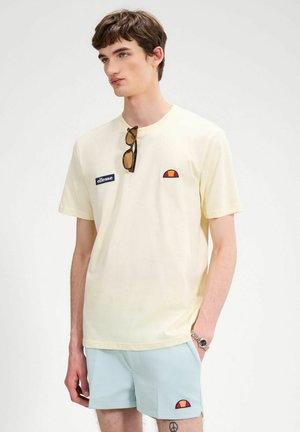 Junger Mann trägt ein cremefarbenes T-Shirt mit Ellesse-Logo, hellblaue Shorts, eine Sonnenbrille hängt am Kragen des T-Shirts und eine goldene Uhr.