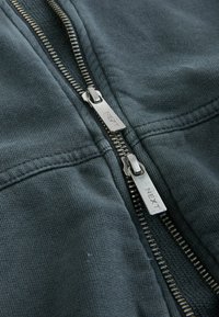 Dunkelgraue Zip-Jacke mit metallischen Reißverschlüssen, die mit "NEXT"-Branding versehen ist. Sie verfügt über einen strukturierten Stoff und sichtbare Nähte entlang der Säume.