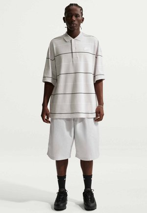 Persona che indossa una polo grigia chiaro oversize con sottili strisce orizzontali, pantaloncini bianchi, calzini Nike neri e scarpe da ginnastica nere, in piedi di fronte.