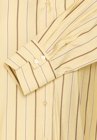 Camicia gialla con bottoni e strisce verticali marroni. Presenta maniche lunghe con polsini e un colletto arrotondato, realizzata in un tessuto morbido.