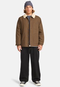 Brown outer jacket with a cream shearling collar, front buttons, two side pockets, paired with black wide-leg pants and sneakers.