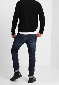 Blå denimjeans med smal passform, medelhög tvätt och uppvikta fållar. Bärs med en svart tröja, som visar bakfickor och sömnadsdetaljer.