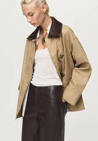 Khaki jacket with a brown collar, featuring a loose fit and button details. Paired with dark brown pants and a white top.