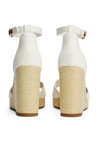 Sandalias de cuña de tela blanca con un tacón de mimbre natural texturizado, tira en el tobillo y diseño de punta abierta. Costuras sutiles en la parte superior de tela.