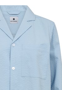 Camisa de pijama azul claro con tejido texturizado, cuello con muesca, un bolsillo en el pecho y cierre de botón. Etiqueta visible dentro del cuello.