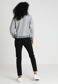 Sudadera gris corta con una banda de logo negra en el dobladillo, combinada con jeans negros y zapatillas blancas. Cabello rizado visible desde atrás.